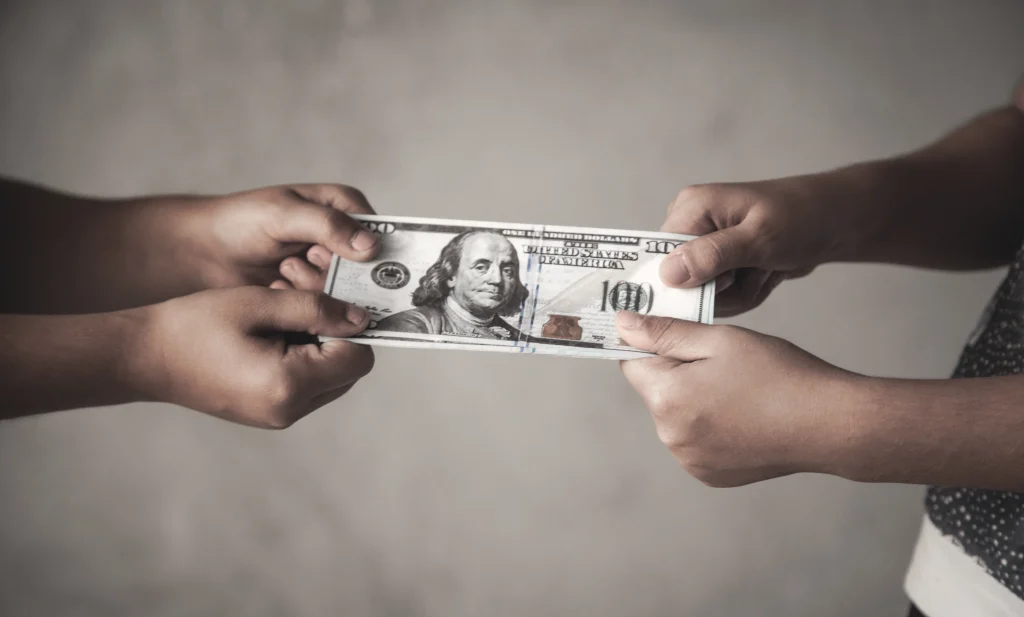 A pair of hands holding a stack of 100 bills on one side and another pair holding it on the other. They are pulling the stack in opposite directions.